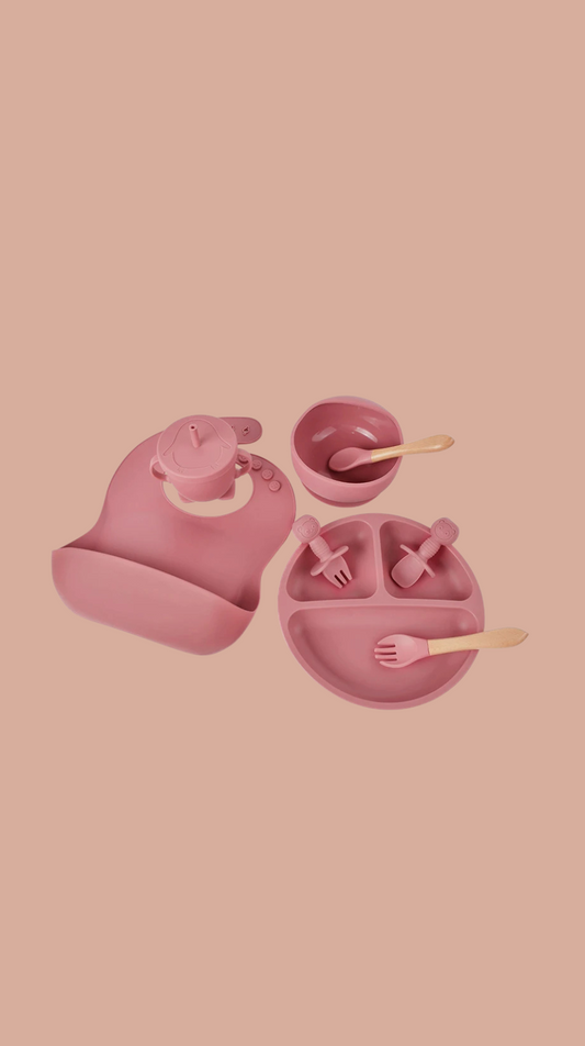 Pink children's dining set including bib, plate, cup, and utensils on a pink background